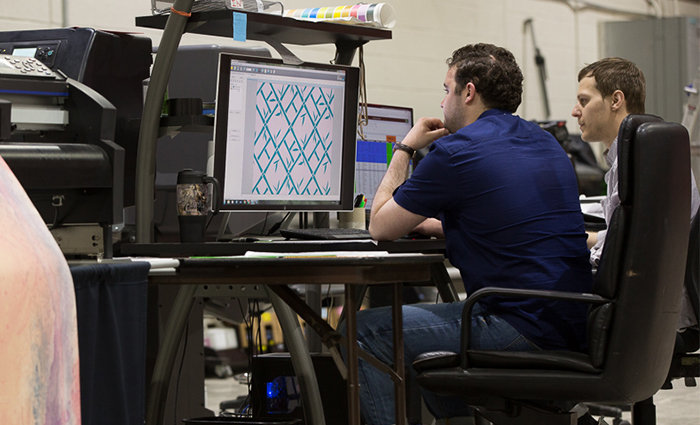 A man sat in a chair at a desk, looking at an incredibly large computer monitor, displaying something complicated.